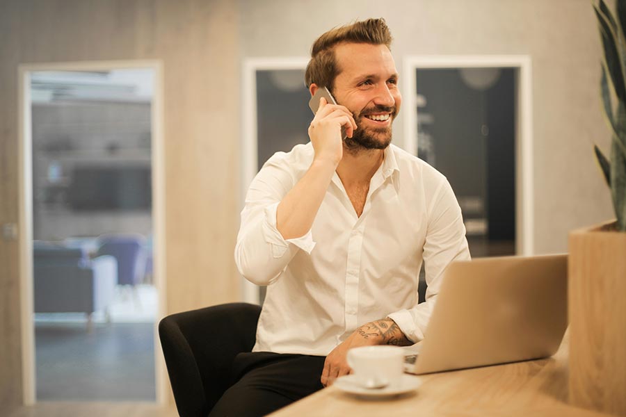Smiling man in white shirt on phone at desk with laptop and coffee
